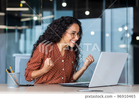 Hispanic business woman celebrating victory success, employee with curly hair inside office reading good news, using laptop at work inside office holding hand up and happy triumph gesture. 96910465