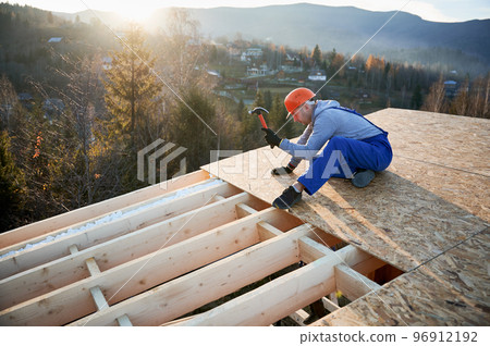 Carpenter hammering nail into OSB panel on the roof top of future cottage at sunrise. Man worker building wooden frame house. Carpentry and construction concept. 96912192