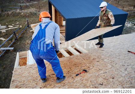 Carpenters mounting wooden OSB panel on rooftop of future cottage. Men workers building wooden frame house in the Scandinavian style barnhouse. Carpentry and construction concept. 96912201