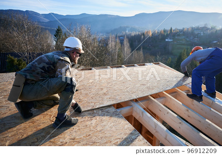 Carpenters mounting wooden OSB board on rooftop of future cottage. Men workers building wooden frame house. Carpentry and construction concept. Carpenters mounting wooden OSB board on rooftop of future cottage. Men workers building wooden frame house. Carpentry and construction concept. 96912209