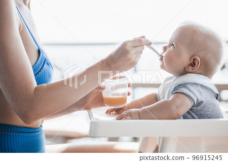 Mother spoon feeding her baby boy child in baby chair with fruit puree on a porch on summer vacations. Baby solid food introduction concept 96915245
