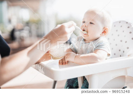 Mother spoon feeding her baby boy child in baby chair with fruit puree on a porch on summer vacations. Baby solid food introduction concept 96915248