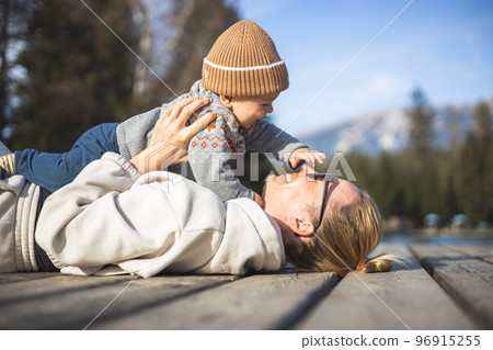 Happy family. Young mother playing with her baby boy infant oudoors on sunny autumn day. Portrait of mom and little son on wooden platform by lake. Positive human emotions, feelings, joy Happy family. Young mother playing with her baby boy infant oudoors on sunny autumn day. Portrait of mom and little son on wooden platform by lake. Positive human emotions, feelings, joy 96915255