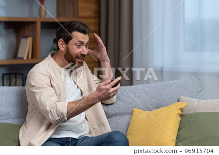 Man at home on sofa using smartphone, celebrating victory in living room reading happy news notifications from phone online holding hand up gesture of triumph and success sitting on sofa. Man at home on sofa using smartphone, celebrating victory in living room reading happy news notifications from phone online holding hand up gesture of triumph and success sitting on sofa. 96915704