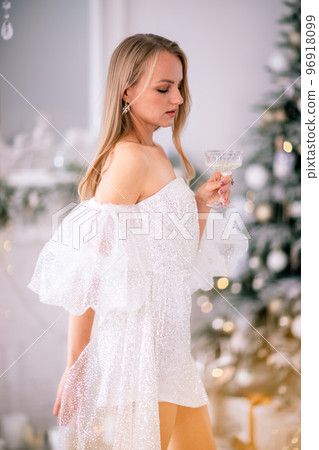 The blonde in the Christmas room. A beautiful blonde woman in a shiny light short dress with a train stands in a beautiful bright room decorated with a festive interior with a Christmas tree. 96918099