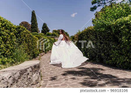 a beautiful woman with long brown hair and long white dress runs along a path along beautiful bushes in the park rear view a beautiful woman with long brown hair and long white dress runs along a path along beautiful bushes in the park rear view 96918106