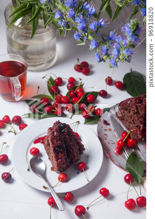 Chocolate cake with sweet cherry, cup of tea and bouquet of blue flowers on white background.. Chocolate cake with sweet cherry, cup of tea and bouquet of blue flowers on white background.. 96918820