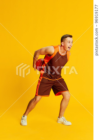 Portrait of young active man in orange uniform training, playing basketball isolated over yellow background. Concept of sport and emotions 96919177