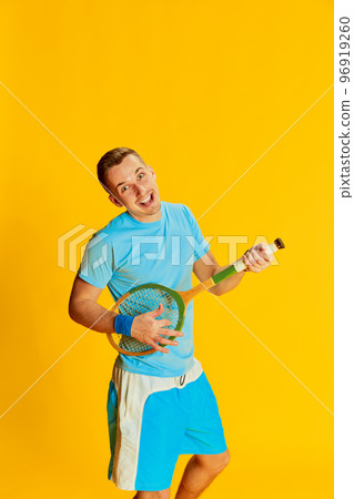 Portrait of young man in blue uniform playing tennis racket like guitar, posing isolated over yellow background. Concept of sport and emotions Portrait of young man in blue uniform playing tennis racket like guitar, posing isolated over yellow background. Concept of sport and emotions 96919260