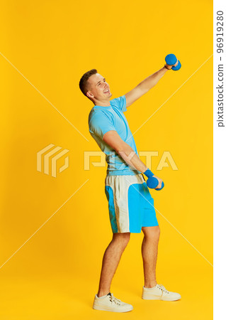 Portrait of young man in blue uniform training, raising hands with dumbbells, posing isolated over yellow background. Concept of sport and emotions 96919280