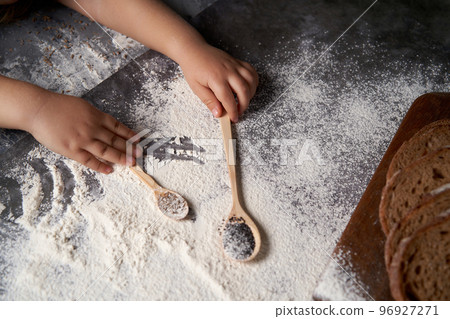 The hands of a kid who helps to make pastries . The child holds wooden spoons with seeds, flour is scattered on the table. High angle. 96927271
