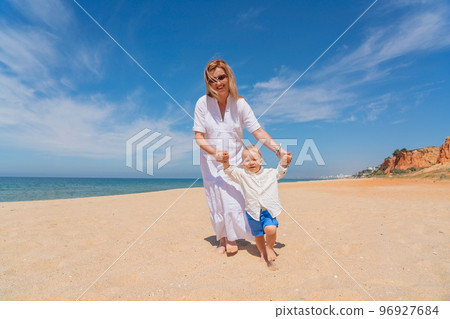 A young caucasian mother walks with her happy son along the sea beach in the summer, taking first steps. Concept of happy childhood and motherhood. 96927684