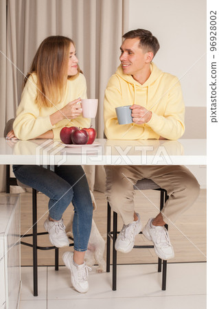 Happy cherrful couple in the kitchen sitting by the table and drinking from the cups Happy cherrful couple in the kitchen sitting by the table and drinking from the cups 96928002