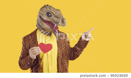 Funny man in dinosaur mask holding red Valentine and pointing at copy space on yellow background 96928680