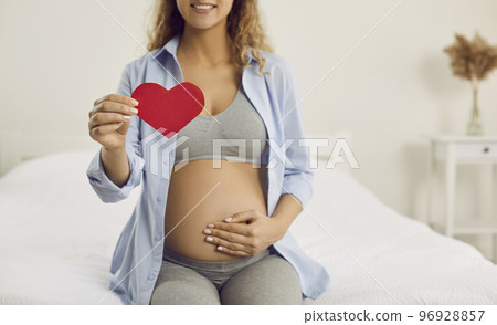 Red paper heart in hand of calm and happy pregnant woman who is in third trimester of pregnancy. Red paper heart in hand of calm and happy pregnant woman who is in third trimester of pregnancy. 96928857