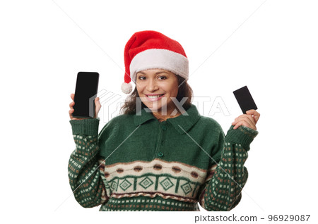Charming multi-ethnic woman wearing Santa hat and warm knitted Xmas patterned sweater, smiling and showing her smartphone and blank credit card with copy space on camera, isolated on white background 96929087