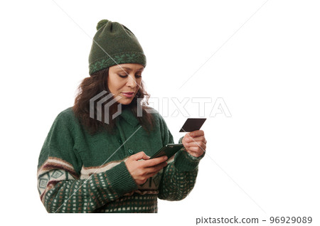 Cheerful Latin American woman wearing Xmas patterned sweater and woolen hat, holding credit card, making online payment while shopping in internet, using her smartphone, isolated over white background 96929089