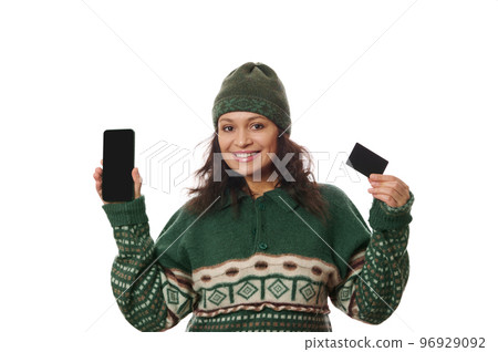Charming multi-ethnic woman wearing warm knitted Xmas patterned sweater and green hat, smiling and showing her smartphone and blank credit card with copy space on camera, isolated on white background 96929092