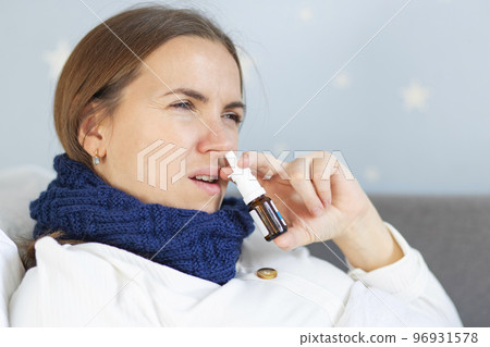 Young woman with bad cold lying in bed under blanket and using nasal drops 96931578