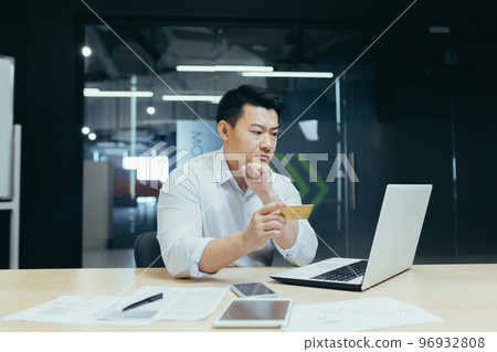 Online shopping, ordering. A young Asian man businessman sits in the office, looks at the laptop and holds a credit card in his hand. Thoughtfully, he makes purchases, places an order, and settles. 96932808