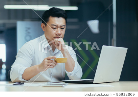 A young man, an Asian businessman, a banker, conducts operations with cards, issuance, registration. Sitting in the office at a table with a laptop, holding and looking at a credit card in his hands. A young man, an Asian businessman, a banker, conducts operations with cards, issuance, registration. Sitting in the office at a table with a laptop, holding and looking at a credit card in his hands. 96932809