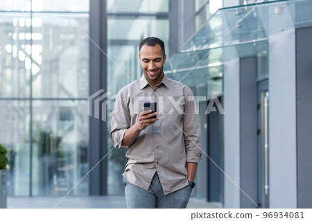 Latin american businessman in casual shirt walking near office building from outside, man smiling and using phone, freelancer typing message and browsing internet pages. 96934081