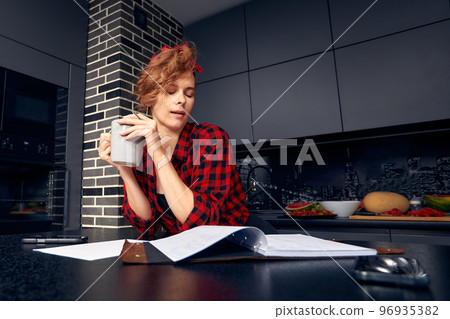Joyful young Hispanic female working from home, doing paperwork, having coffee break, sitting at dining table with mug and diary, making to do list. Cute girl sketching in copybook and drinking tea 96935382