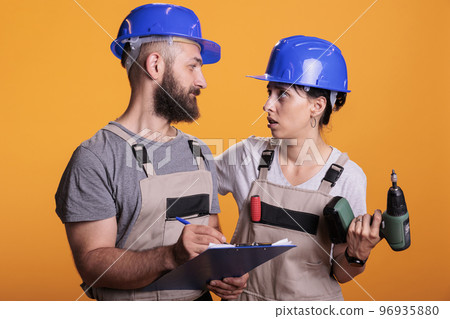 Construction experts taking notes on clipboard and checking renovation measurement, holding drill gun. Man and woman renovators looking at papers or files, standing over yellow background. Construction experts taking notes on clipboard and checking renovation measurement, holding drill gun. Man and woman renovators looking at papers or files, standing over yellow background. 96935880