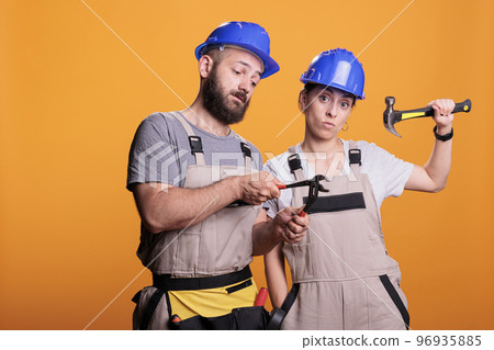 Confident constructors holding renovating tools on camera, using sledgehammer and pair of pliers to work on renovation project. Builders team posing with hammer and pliers over background. Confident constructors holding renovating tools on camera, using sledgehammer and pair of pliers to work on renovation project. Builders team posing with hammer and pliers over background. 96935885
