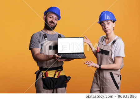 Construction workers holding laptop with white display, wearing uniforms and helmets. Showing empty blank screen with isolated copyspace on mockup template, using portable computer. Construction workers holding laptop with white display, wearing uniforms and helmets. Showing empty blank screen with isolated copyspace on mockup template, using portable computer. 96935886