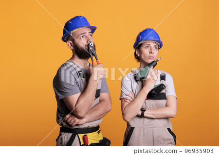 Team of builders brainstorming construction ideas in studio, thinking about renovating solution to start rebuilding work. Pensive man and woman holding power drilling gun and pair of pliers. Team of builders brainstorming construction ideas in studio, thinking about renovating solution to start rebuilding work. Pensive man and woman holding power drilling gun and pair of pliers. 96935903