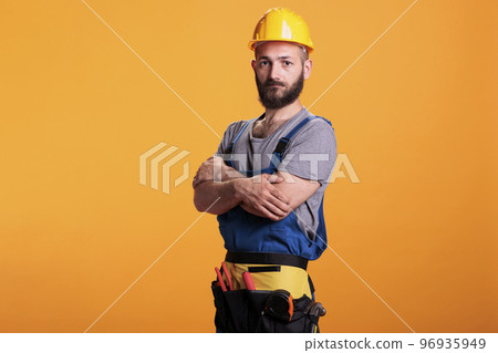 Cheerful repairman carpenter with renovating uniform and hardhat posing on camera in studio. Professional handyman preparing to work on refurbishment project, craftsman expert. 96935949