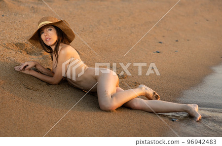 Naked young brunette in hat lying on wet sandy shore by ocean Naked young brunette in hat lying on wet sandy shore by ocean 96942483