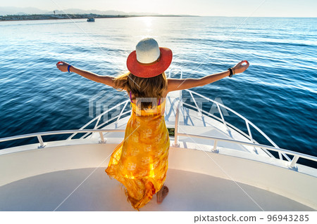 Young woman with long hair wearing yellow dress and straw hat standing with raised hands on white yacht deck enjoying view of blue sea water. Young woman with long hair wearing yellow dress and straw hat standing with raised hands on white yacht deck enjoying view of blue sea water. 96943285