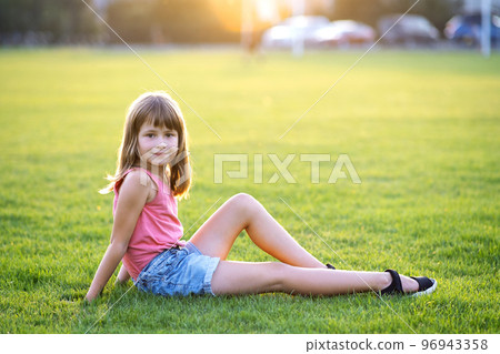 Young pretty child girl sitting on fresh green grass lawn on warm summer day outdoors. 96943358