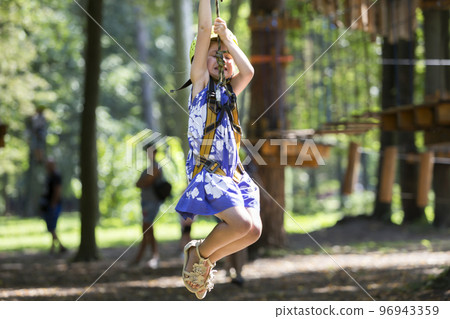 Young pretty blond child girl in safety harness and helmet on rope way on bright bokeh background. 96943359