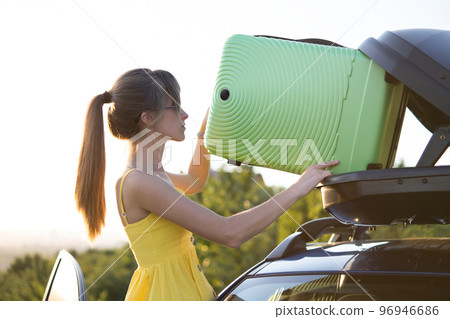 Female driver in summer dress putting green suitcase inside her car roof rack. Travel and vacations concept. 96946686