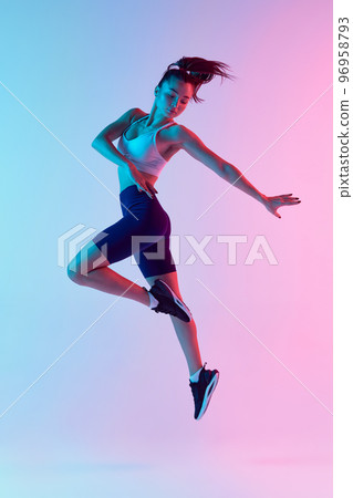 Portrait of young sportive woman training, doing exercises, jumping isolated over gradient blue pink background in neon light. Concept of sport, fitness, health 96958793