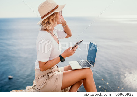 Digital nomad, Business woman working on laptop by the sea. Pretty lady typing on computer by the sea at sunset, makes a business transaction online from a distance. Freelance, remote work on vacation 96960922