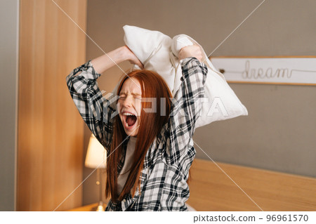 Close-up of angry irritated annoyed woman shouting and yelling with aggressive expression, throwing pillow sitting on bed in bedroom. Furious crazy redhead female vented anger in privacy of home. Close-up of angry irritated annoyed woman shouting and yelling with aggressive expression, throwing pillow sitting on bed in bedroom. Furious crazy redhead female vented anger in privacy of home. 96961570