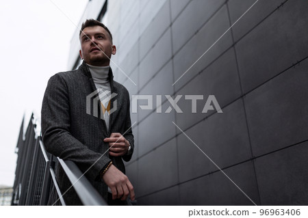 brutal stylish man in a gray coat on the stairs against a gray wall brutal stylish man in a gray coat on the stairs against a gray wall 96963406