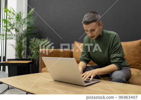 Strong woman with short haircut working on laptop while sitting on sofa at home Strong woman with short haircut working on laptop while sitting on sofa at home 96963407