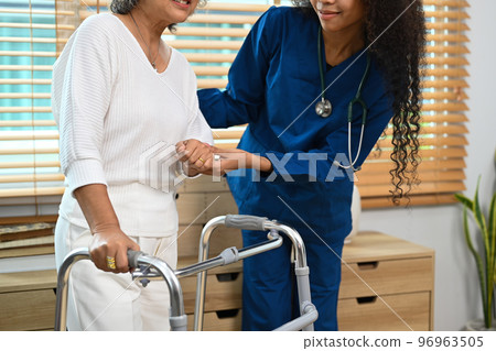Young caregiver assisting, teaching senior woman to walk with walker. Assistance, rehabilitation and health 96963505