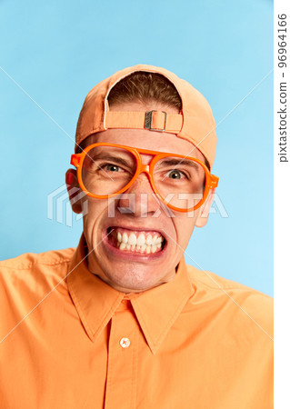 Closeup portrait of emotional young man in eyewear and cap looking at camera with vivid emotions isolated over light blue background. Mental health, facial expression, emotions concept Closeup portrait of emotional young man in eyewear and cap looking at camera with vivid emotions isolated over light blue background. Mental health, facial expression, emotions concept 96964166