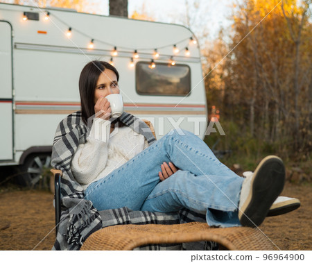 Caucasian woman in a knitted hat wrapped in a plaid and drinks a warming drink outdoors. Travel in a motor home in the fall. 96964900