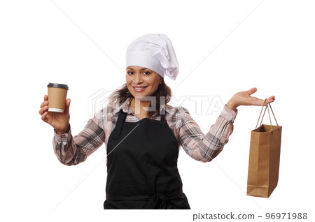 Pleasant Latin American woman barkeeper, waitress wearing chef apron and hat, holding takeaway hot drink in disposable cardboard mug and recyclable paper bag. Food consumerism and delivery concept Pleasant Latin American woman barkeeper, waitress wearing chef apron and hat, holding takeaway hot drink in disposable cardboard mug and recyclable paper bag. Food consumerism and delivery concept 96971988