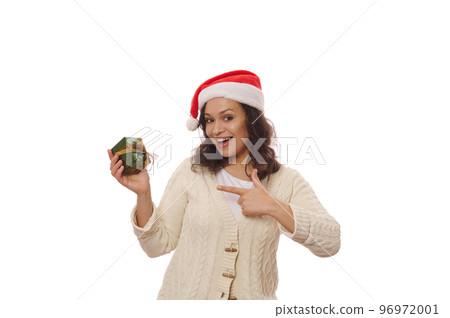 African American pretty woman in Santa hat and beige sweater, pointing at a happy Christmas present, smiling looking at camera, isolated over white background. Boxing Day. Time to open gift boxes 96972001