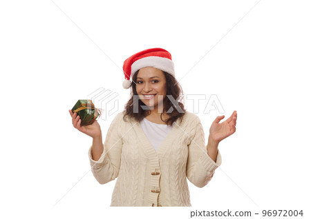 Attractive woman wearing Santa hat, holding Christmas present, smiling a cheerful toothy smile looking at camera, isolated on white background. Copy ad space. Festive mood of upcoming winter holidays 96972004