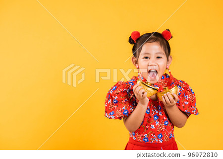 Chinese New Year. Happy Asian Chinese little girl smile wearing red cheongsam holding gold ingot, Portrait children in traditional dress hold golden bar, studio short isolated on yellow background Chinese New Year. Happy Asian Chinese little girl smile wearing red cheongsam holding gold ingot, Portrait children in traditional dress hold golden bar, studio short isolated on yellow background 96972810