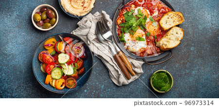 Middle Eastern breakfast or brunch with Shakshouka in pan, toasts, vegetables salad, hummus, olives 96973314